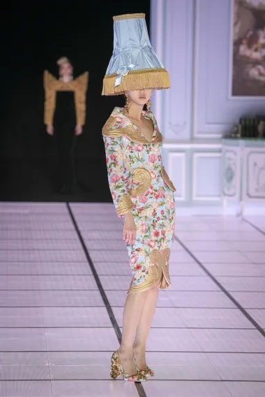 Suknja i sako cvjetnog uzorka sa zlatnim detaljima u kombinaciji s cipelama sličnog uzorka neobična su kombinacija sami po sebi, no Moschino je outfit dodatno &quot;začinio&quot; plavo-zlatnom lampom na glavi modela koja mu prekriva oči. Malo bizarno, zar ne?