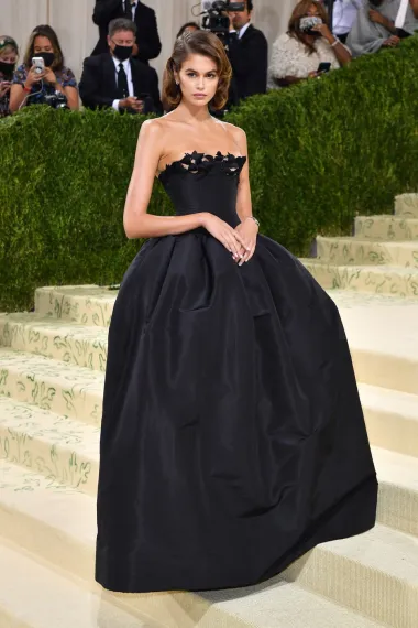 Kaia Gerber nosila je elegantnu crnu balsku haljinu brenda Oscar de la Renta, nadahnutu jednom od najpoznatijih modnih kombinacija ikone stila — Priscille Presley s Met Gale 1981. Haljina bez naramenica, s naglašenim korzetom i voluminoznom suknjom, odavala je dojam klasičnog stila. 
